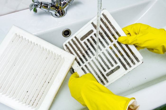 Une personne portant des gants en caoutchouc jaune rince le couvercle et le filtre d'un ventilateur d'extraction de salle de bain dans l'évier.