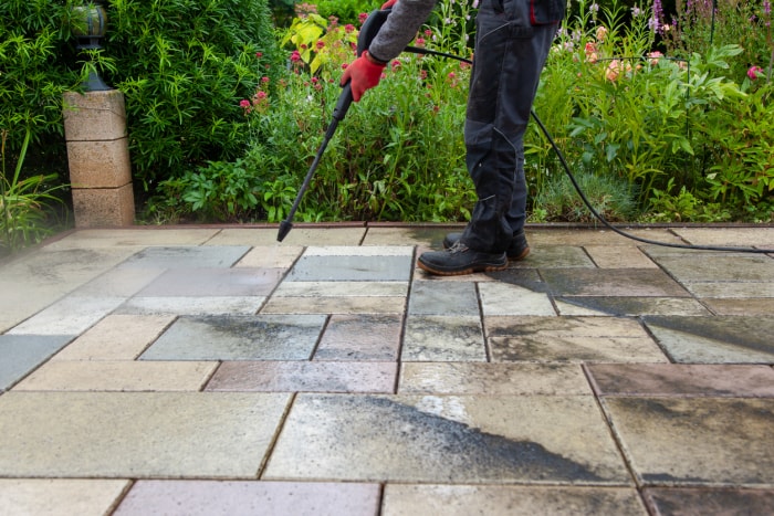 un homme, de la taille aux pieds, lave au jet d'eau les dalles d'une terrasse près des fleurs