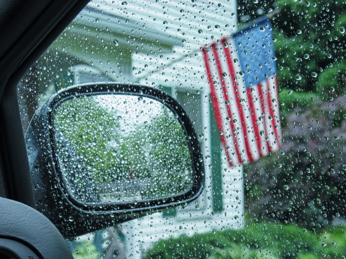 vue de l'intérieur d'une voiture d'un drapeau américain suspendu à une maison avec les fenêtres et le rétroviseur couverts de gouttes de pluie