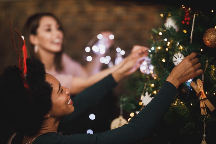 Une femme afro-américaine heureuse profite des vacances à la maison, décore son sapin de Noël et arrange les lumières de Noël avec des amis.