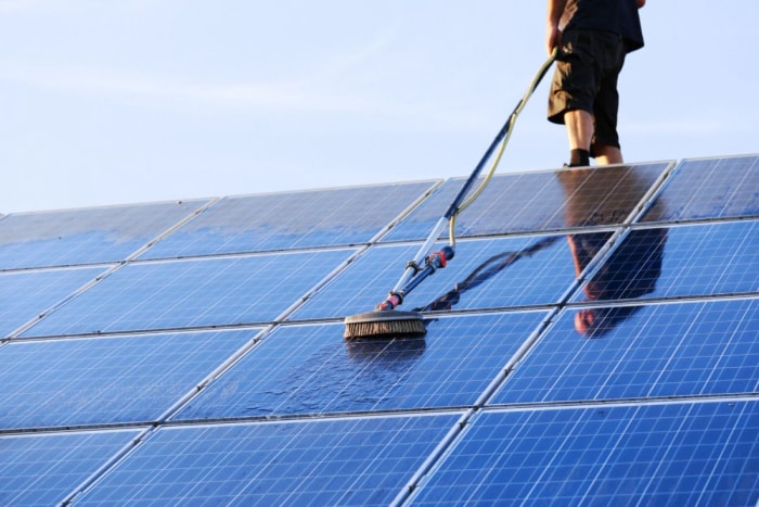 Un ouvrier, dont on voit le bas du corps, nettoie un système de panneaux solaires sur un toit à l'aide d'une brosse et d'un tuyau d'arrosage.