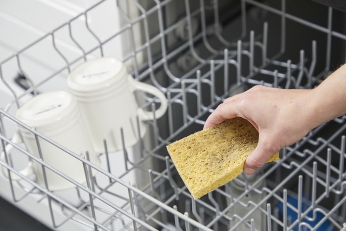 Une femme met une éponge jaune dans le panier supérieur du lave-vaisselle avec des tasses.