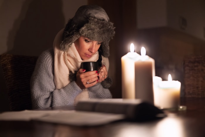 Une jeune femme gelée en vêtements d'hiver se réchauffe les mains avec une tasse de thé et s'allume avec des bougies alors que les pannes d'électricité provoquent des pannes de courant.