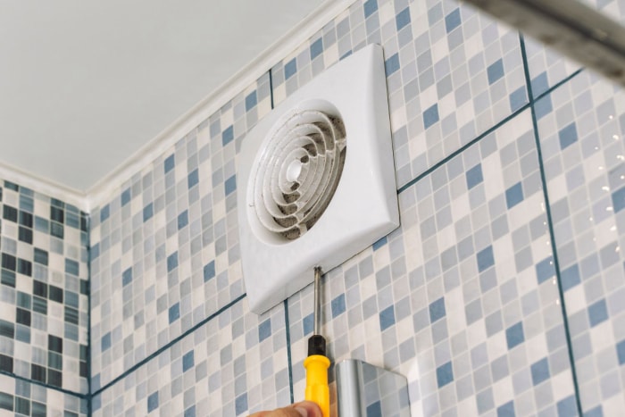 Une personne dont seule une partie de la main est visible utilise un tournevis pour retirer ou installer un ventilateur d'extraction sur un mur de salle de bain carrelé.