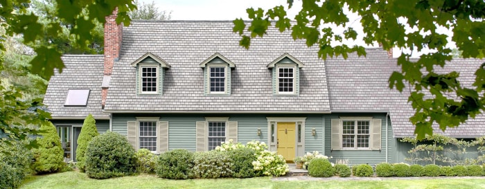 Une maison à deux étages avec des lucarnes est peinte en bleu pâle avec une porte jaune et de la verdure autour.