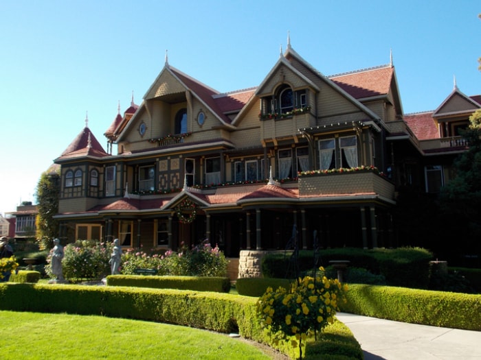 une maison victorienne avec une variété de pignons et de tuiles rouges sur un extérieur beige se trouve derrière des jardins formels contre un ciel bleu