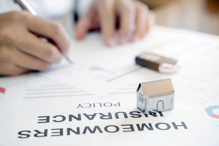 Une petite maquette de maison est posée sur un document intitulé « Police d'assurance habitation » tandis qu'une personne écrit en arrière-plan.