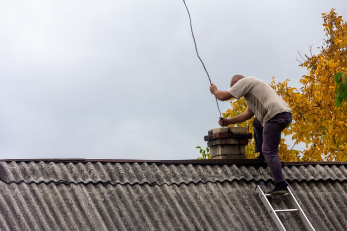 un ramoneur nettoie la cheminée de la suie sur le toit d'une maison de village contre un ciel gris avec un espace de copie, se préparant pour la saison de chauffage