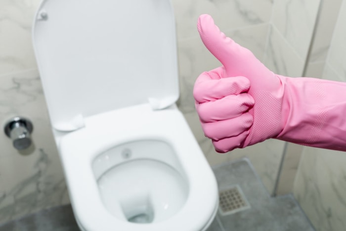 Une main gantée de rose faisant le signe du pouce levé devant des toilettes propres avec le couvercle relevé.