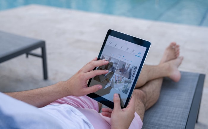 Une personne se prélassant au bord d'une piscine accède aux images des caméras de sécurité visionnées sur une tablette. 