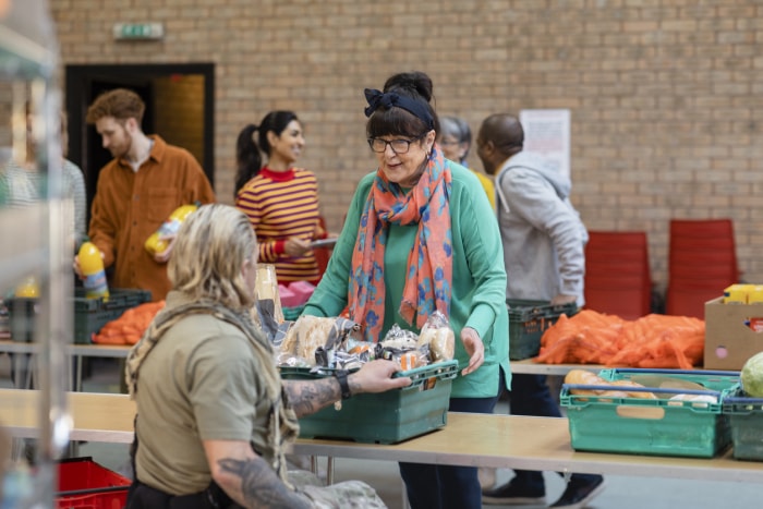 une femme avec des lunettes et un foulard orange vif passe une caisse de nourriture à un autre bénévole sur une longue table dans une banque alimentaire avec des bénévoles qui trient la nourriture sur les tables.