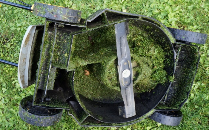 Une tondeuse à gazon électrique est à l'envers sur une pelouse, montrant de l'herbe mouillée obstruant les lames.