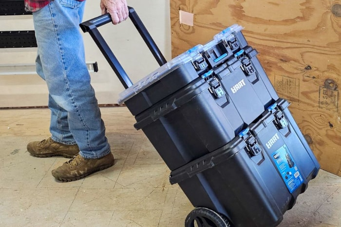 Une personne tient la poignée télescopique de la boîte à outils du système d'empilage Hart Tools tout en la faisant rouler dans une salle de travail.