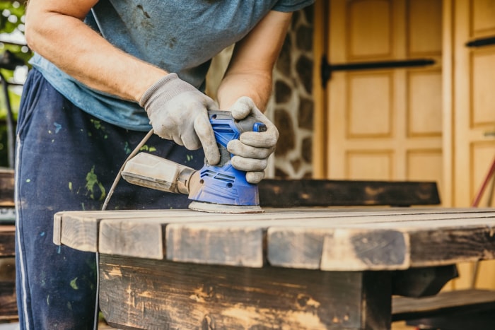 Une personne ponce l'ancienne finition d'une table d'extérieur pour la préparer à recevoir une nouvelle couche de peinture.