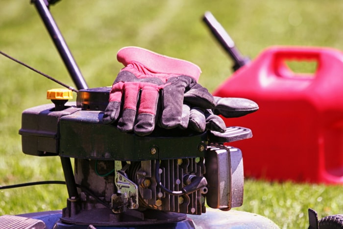 Une paire de gants de jardinage est posée sur une tondeuse à gazon et un bidon d'essence est posé sur l'herbe derrière la tondeuse.