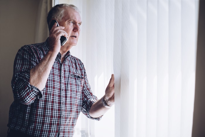 un homme âgé regarde par la fenêtre en écartant les stores et parle au téléphone portable