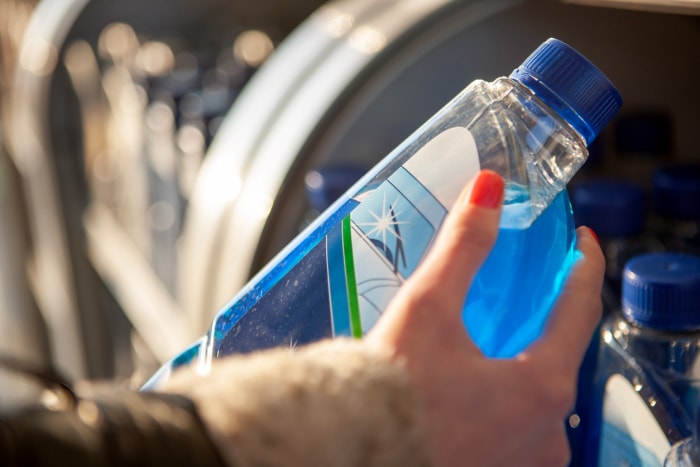 main d'une femme avec un ongle rouge tenant une bouteille de liquide lave-glace bleu devant un moteur de voiture ouvert