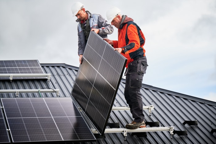 Deux hommes portant des casques de sécurité installent un panneau solaire sur le toit d'une maison.