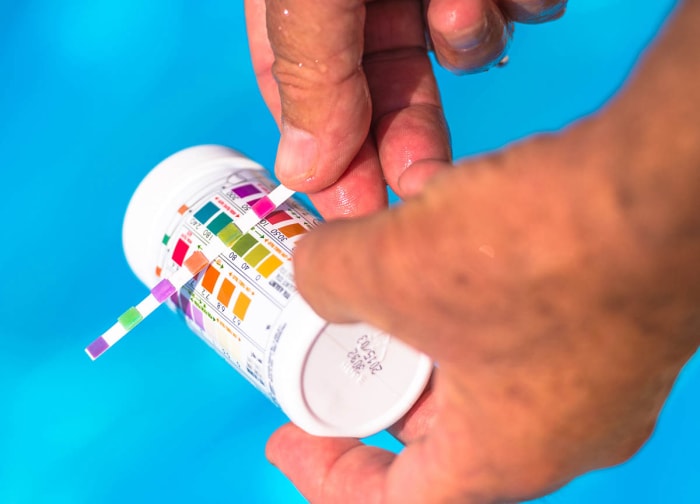 Une personne compare une bandelette de test de piscine à la bouteille tout en testant l’eau d’une piscine.