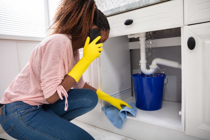 a-woman-in-pink-shirt-and-jeans-wears-yellow-rubber-gloves-while making-a-phone-call-and looking-at-a-leak-under-the-sink-caught-by-a-bucket (une femme en chemise rose et en jean porte des gants en caoutchouc jaune tout en téléphonant et en regardant une fuite sous l'évier, attrapée par un seau)