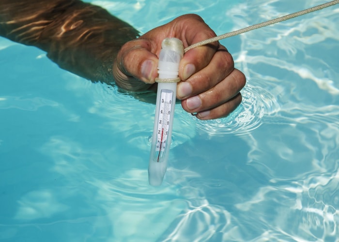 Une main tenant un thermomètre dans une piscine chauffée par la meilleure option de chauffage de piscine.