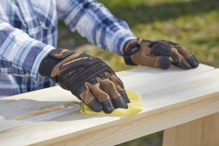 Une femme portant des gants de travail essuie du bois poncé avec un chiffon jaune.