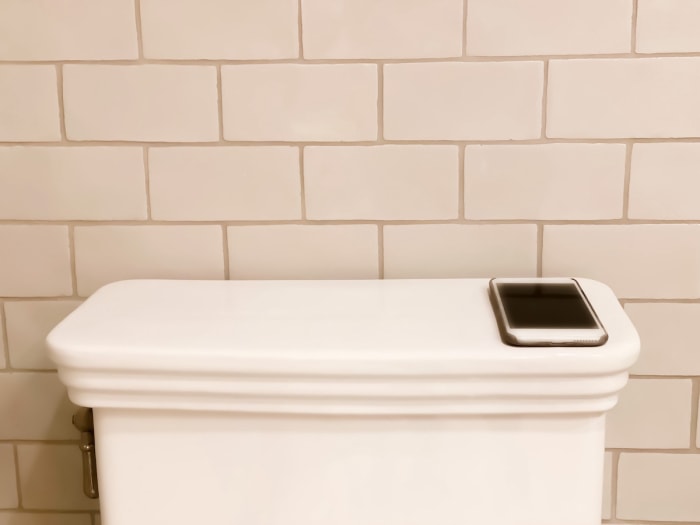 Téléphone intelligent posé à l'arrière d'un réservoir de toilettes blanc avec des carreaux de métro blancs en arrière-plan.