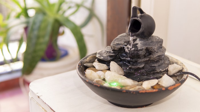 Fontaine zen de table en pierre foncée avec des pierres à l'intérieur, avec une plante en arrière-plan.