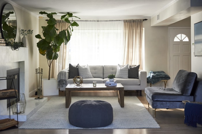 Le salon d'une maison de banlieue comprenant un canapé, un fauteuil et un pouf est éclairé par la lumière du soleil provenant d'une grande fenêtre.