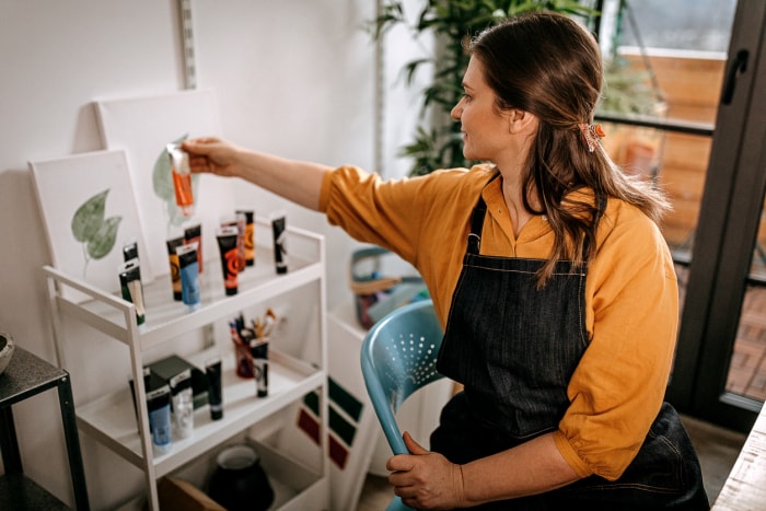 Une femme en chemisier jaune et tablier noir organise des peintures sur un chariot à plusieurs niveaux.