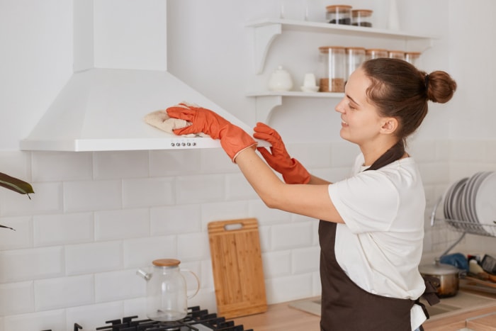 Plan horizontal d'une femme lavant avec un chiffon une hotte de nettoyage électrique dans la cuisine de sa maison, portant un t-shirt blanc, un tablier marron et des gants orange