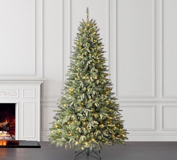 Le sapin de Noël Brighton Spruce Prelit de Holiday Living est illuminé de lumières blanches à côté d'un mur blanc et d'une cheminée blanche avec un feu qui brûle.