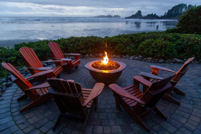 Des chaises Adirondack rouges sont disposées en cercle autour d'un foyer sur une terrasse au bord d'un lac.