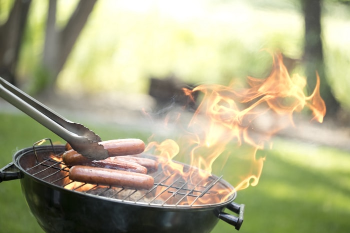 gril rond à l'extérieur sur une pelouse verte avec des pinces plaçant des hot-dogs sur une flamme nue élevée