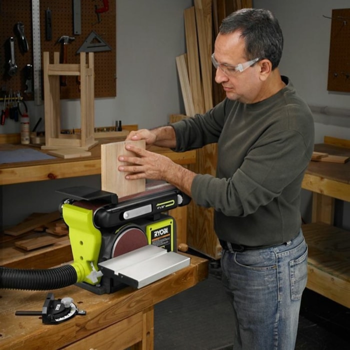 Homme avec des lunettes ponçant du bois avec une grande ponceuse de table verte.