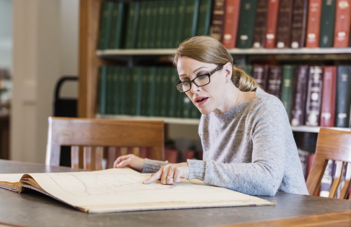 Une femme mature dans la quarantaine faisant des recherches à la bibliothèque, assise à une table et regardant un grand vieux livre.