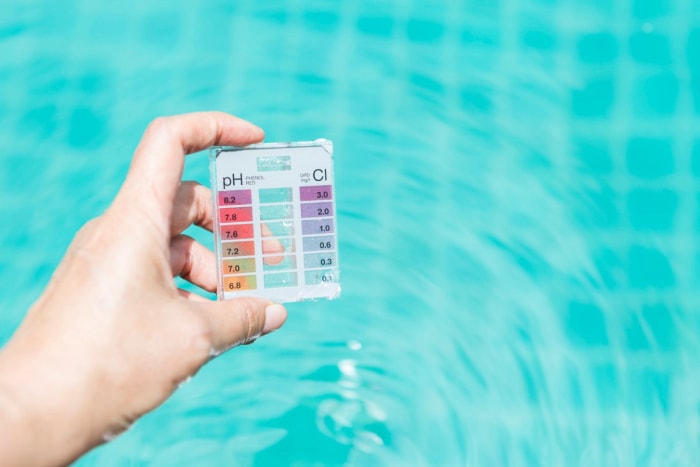 Une personne tient un petit kit de test de l'eau de piscine avec un tableau en couleur pour l'équilibre du pH et les lectures du niveau de chlore imprimé sur le devant.