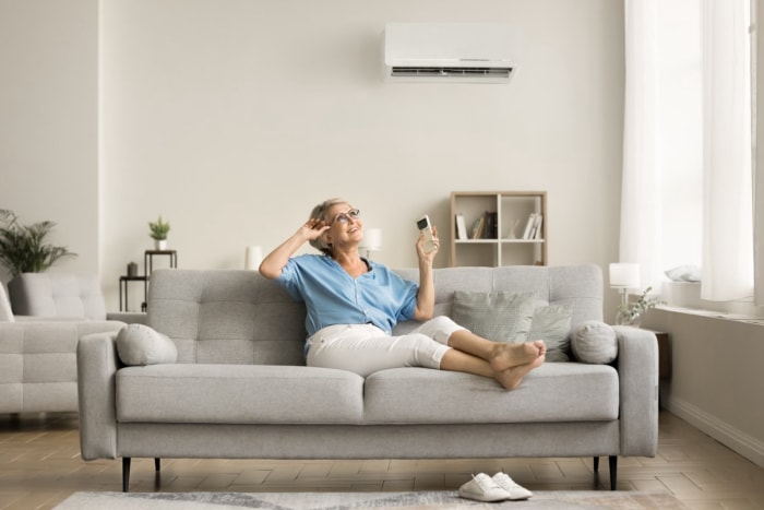 Une femme est assise sur un canapé gris et tient une télécommande qui contrôle la climatisation installée sur le mur blanc derrière elle. 