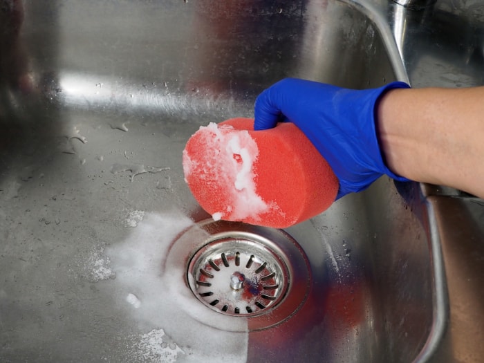 Une personne portant des gants bleus utilise une éponge rouge, du bicarbonate de soude et du vinaigre pour frotter l'évier en acier inoxydable.