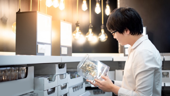 jeune-personne-en-chemise-blanche-regarde-un-luminaire-dans-une-allée-dans-un-magasin-de-détail-avec-d'autres-luminaires-en-exposition-et-dans-des-boîtes-en-étagère