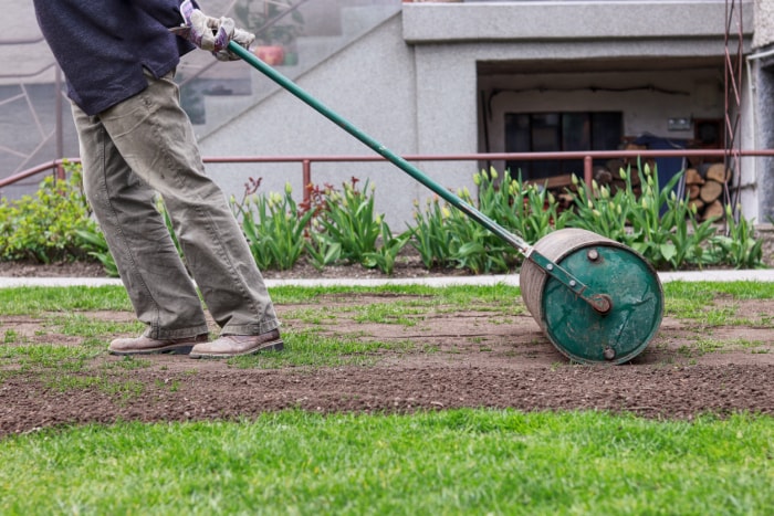 Un homme en tenue de travail tire un rouleau à gazon derrière lui. Nécessité après un long hiver et un long printemps pour une surface plane. Aménagement paysager du jardin. Travailleur d'été