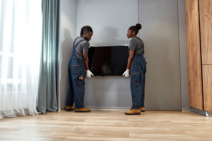 Deux ouvriers en bleu de travail tiennent une télévision contre un mur gris, comme s'ils avaient l'intention de la monter.