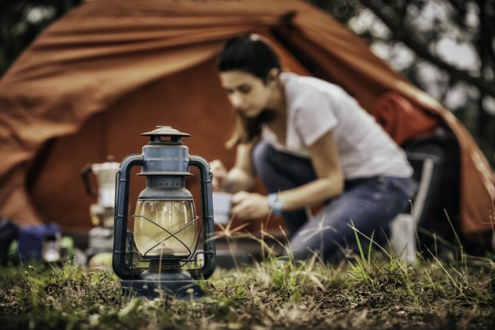 lanterne en gros plan devant un camping où une femme a installé une tente rouge et est floue en arrière-plan en train de faire un feu