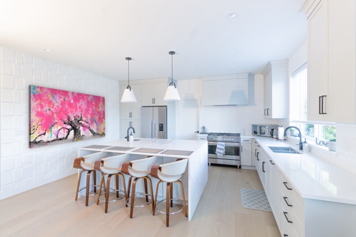 Grande cuisine blanche spacieuse avec une péninsule en marbre et des tabourets de bar en bois, une cuisinière et un réfrigérateur en acier inoxydable, un tableau mural et deux lampes suspendues.