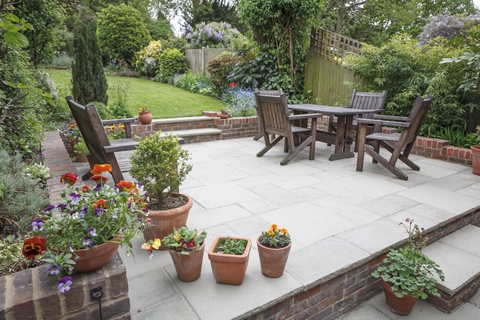 Un jardin aménagé avec un patio en pierre et en brique, une table et des chaises en bois, et des fleurs en pot.