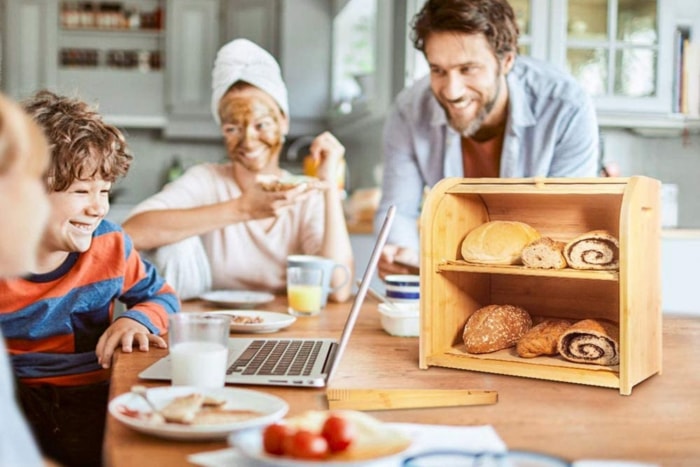 Une famille est réunie devant un comptoir de cuisine avec un ordinateur portable, des produits pour le petit-déjeuner et une boîte à pain remplie de pain.