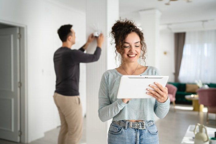 Une femme sourit tout en utilisant une tablette intelligente ; un homme derrière elle installe un système de sécurité domestique. 