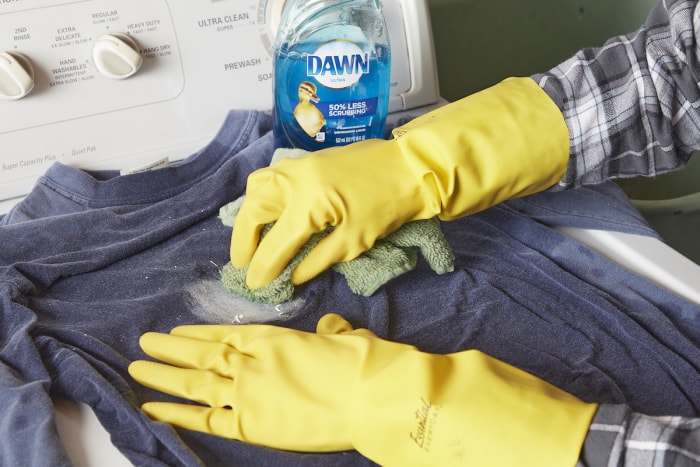 Une femme portant des gants en caoutchouc utilise du savon à vaisselle et un chiffon pour enlever une tache de peinture sur une chemise bleue.