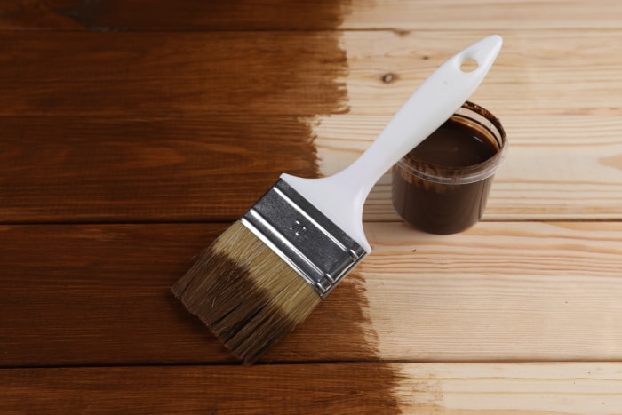 Un pinceau humide et un récipient en plastique contenant de la teinture pour bois brun foncé sont posés sur une table en bois à moitié teinte.