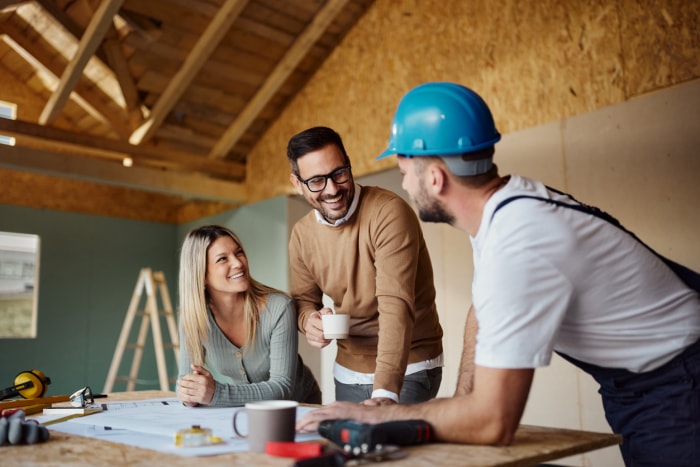 Un jeune couple s'entretient avec un ouvrier du bâtiment portant un casque dans une zone de construction au sujet de plans.